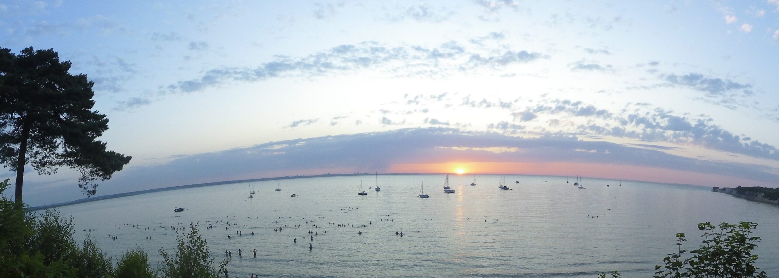 Wide angle shot of Studland Bay with lots of boats on the water and the sun rising on the horizon.