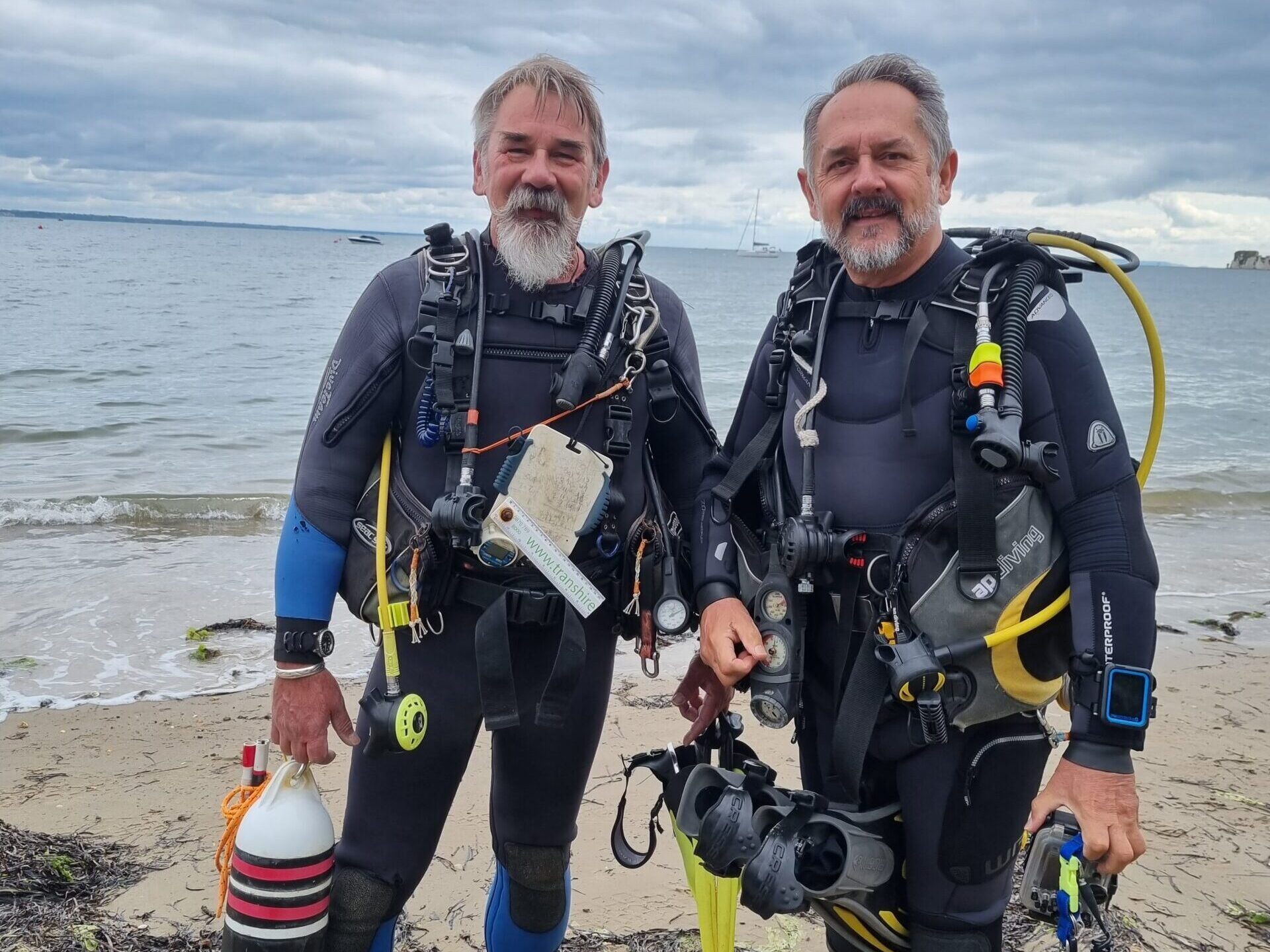 Two divers in full kit standing on the shoreline.