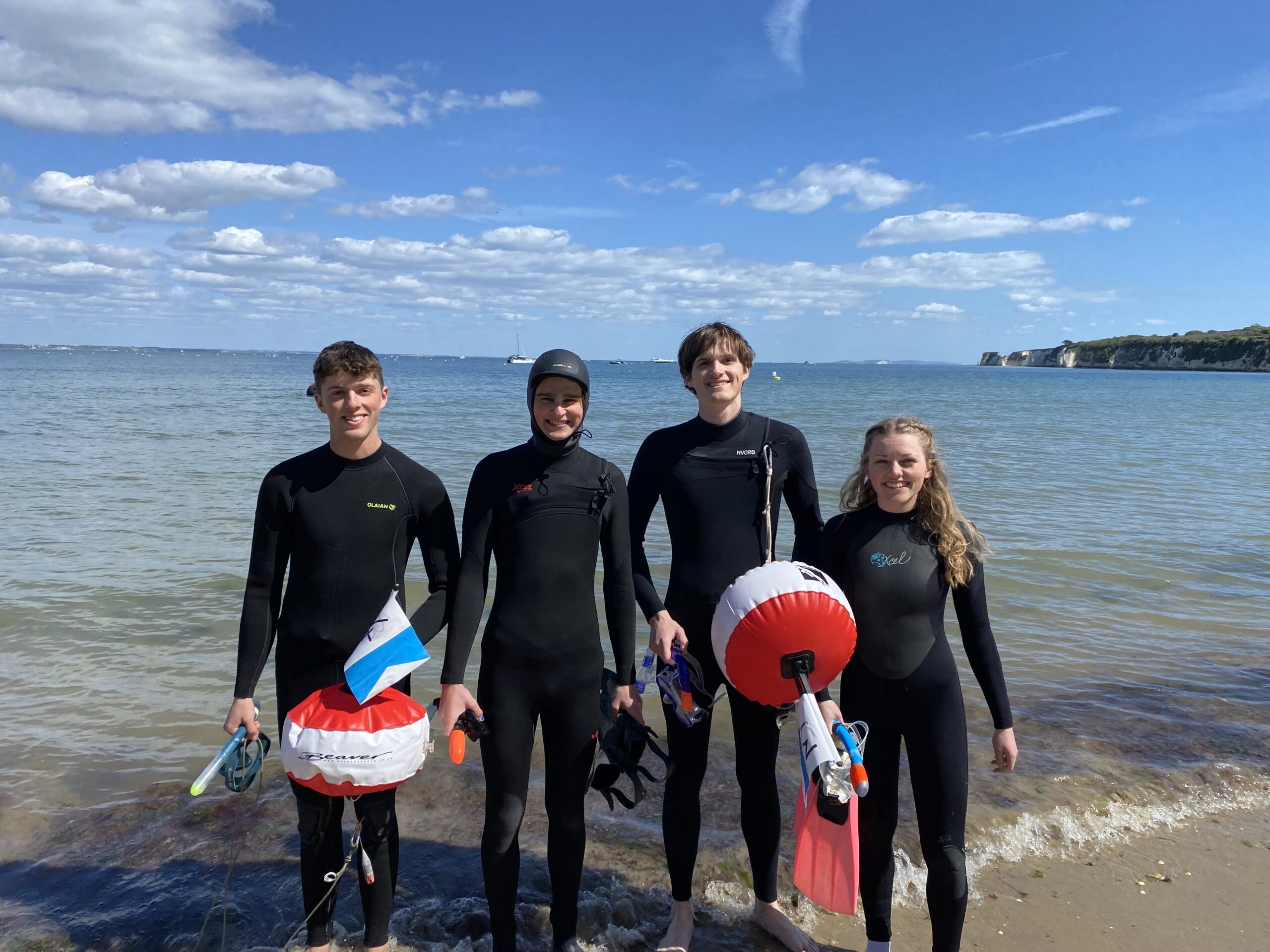 Four students in wetsuits holding diving buoys, standing on the beach with sea and views of Old Harry Rocks behind them.