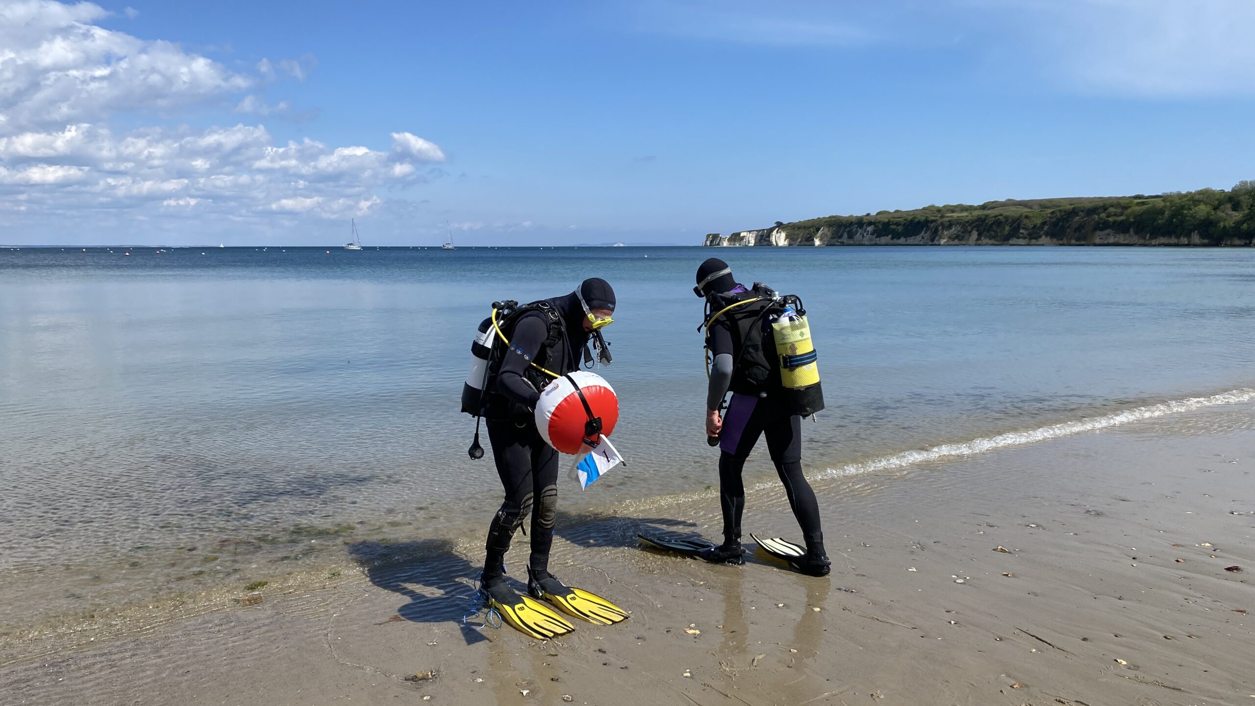 Two students in full diving gear standing by the shore with views of the sea and Old Harry Rocks behind.