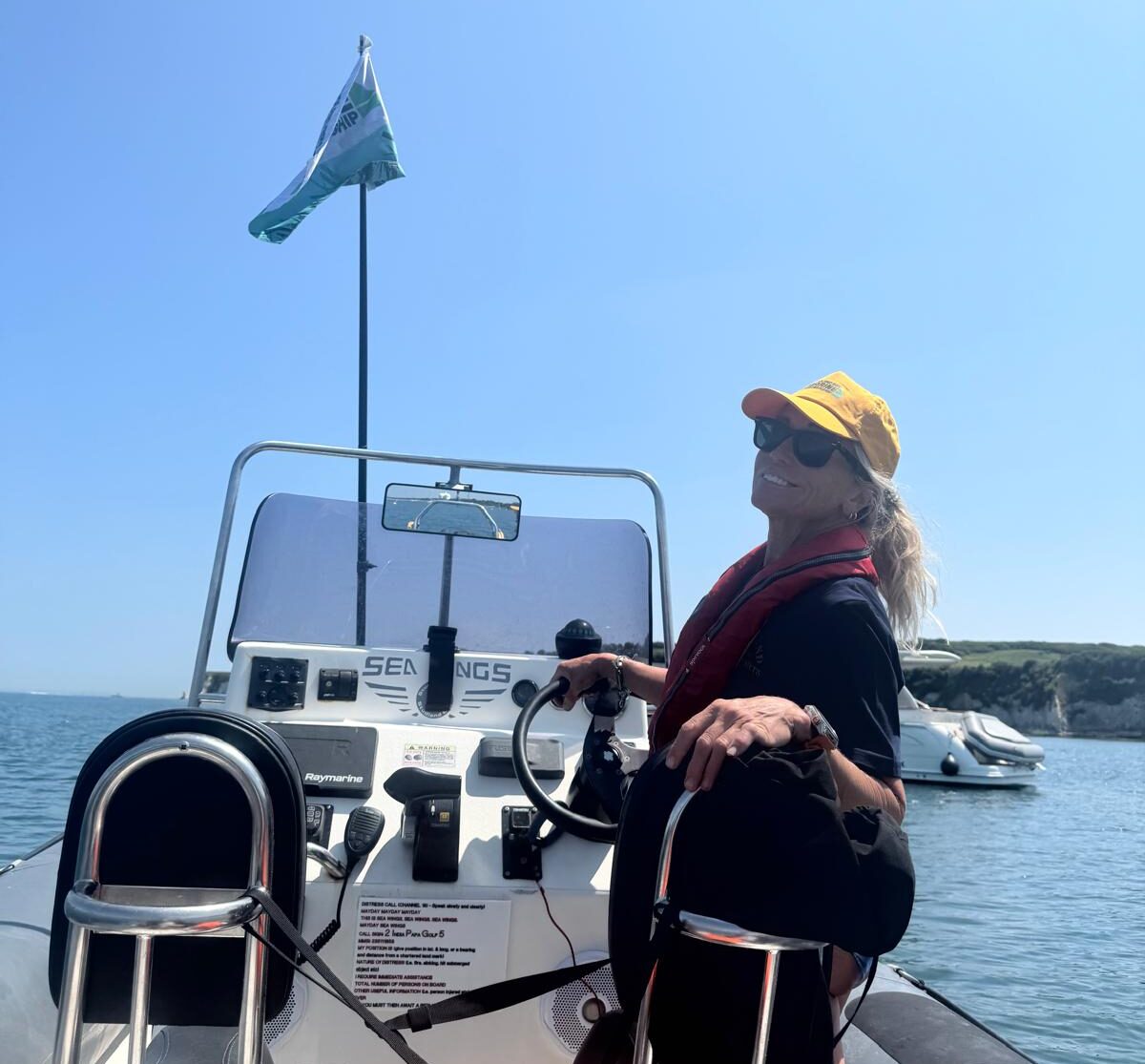 Woman at the helm of a speedboat, wearing a yellow cap. Beyond is the sea and a white boat.